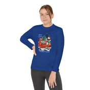 A person is wearing an Urban SoulShine & co Here Comes Donkey Claus Long Sleeve Competitor Tee (Youth Unisex) in blue, showcasing a festive design with a red car, tree, and the phrase "Here Comes Donkey Claus." The athletic fit enhances their style as they stand confidently with one hand on their hip. Their long hair is tied back, and they complete the look with dark pants.