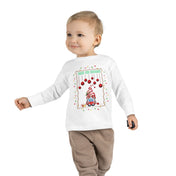 A small child walks confidently in the "Deck The Gnomes Christmas Toddler Long Sleeve Tee" by Urban SoulShine & co, made of 100% cotton. This white tee showcases a festive holiday gnome design beneath red ornaments, accented with green text reading "Peek the Gnome." It pairs perfectly with light brown pants.