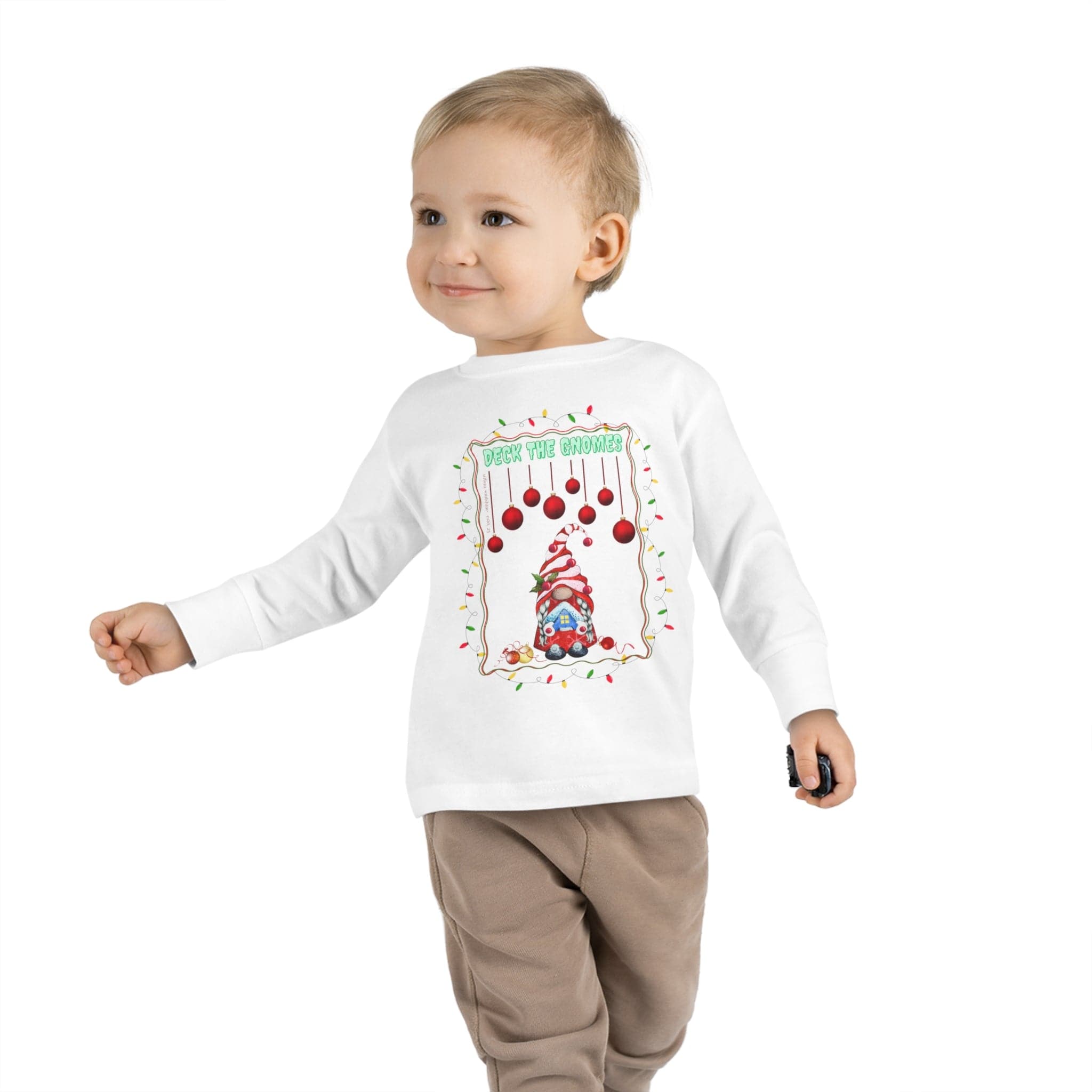 A small child walks confidently in the "Deck The Gnomes Christmas Toddler Long Sleeve Tee" by Urban SoulShine & co, made of 100% cotton. This white tee showcases a festive holiday gnome design beneath red ornaments, accented with green text reading "Peek the Gnome." It pairs perfectly with light brown pants.