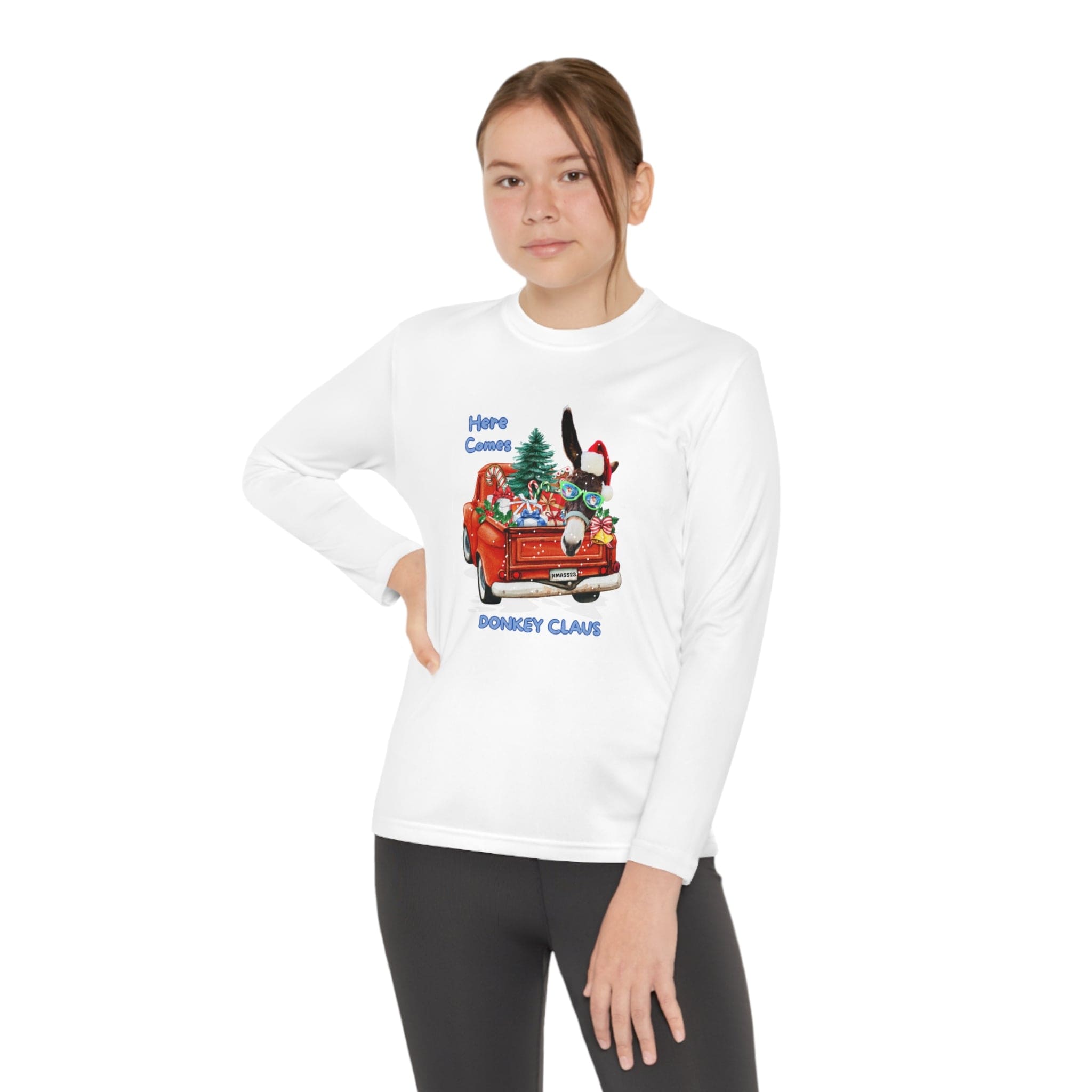 Wearing an athletic-fit shirt from Urban SoulShine & co, the person confidently poses against a plain backdrop. The white long-sleeve tee, named "Here Comes Donkey Claus Long Sleeve Competitor Tee (Youth Unisex)," showcases a festive graphic of a red car filled with presents, a donkey, and a Christmas tree. It is crafted from moisture-wicking polyester and features the playful text "Here Comes Donkey Claus.