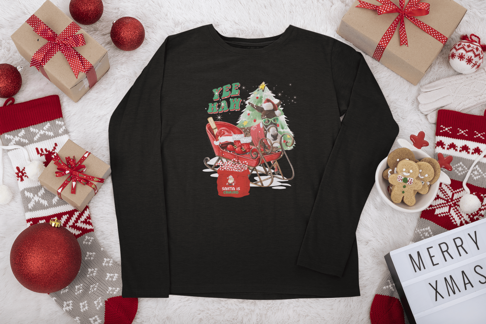 A YEE HAW Donkey Claus Long Sleeve Competitor Tee (Youth Unisex) by Urban SoulShine & co, showcasing an athletic fit and festive design with a red sleigh, Christmas tree, and "YEE HAW" text, lies on a fluffy white surface. Enhanced with moisture-wicking technology, it is surrounded by wrapped gifts, red and white stockings, ornaments, and a "MERRY XMAS" sign.