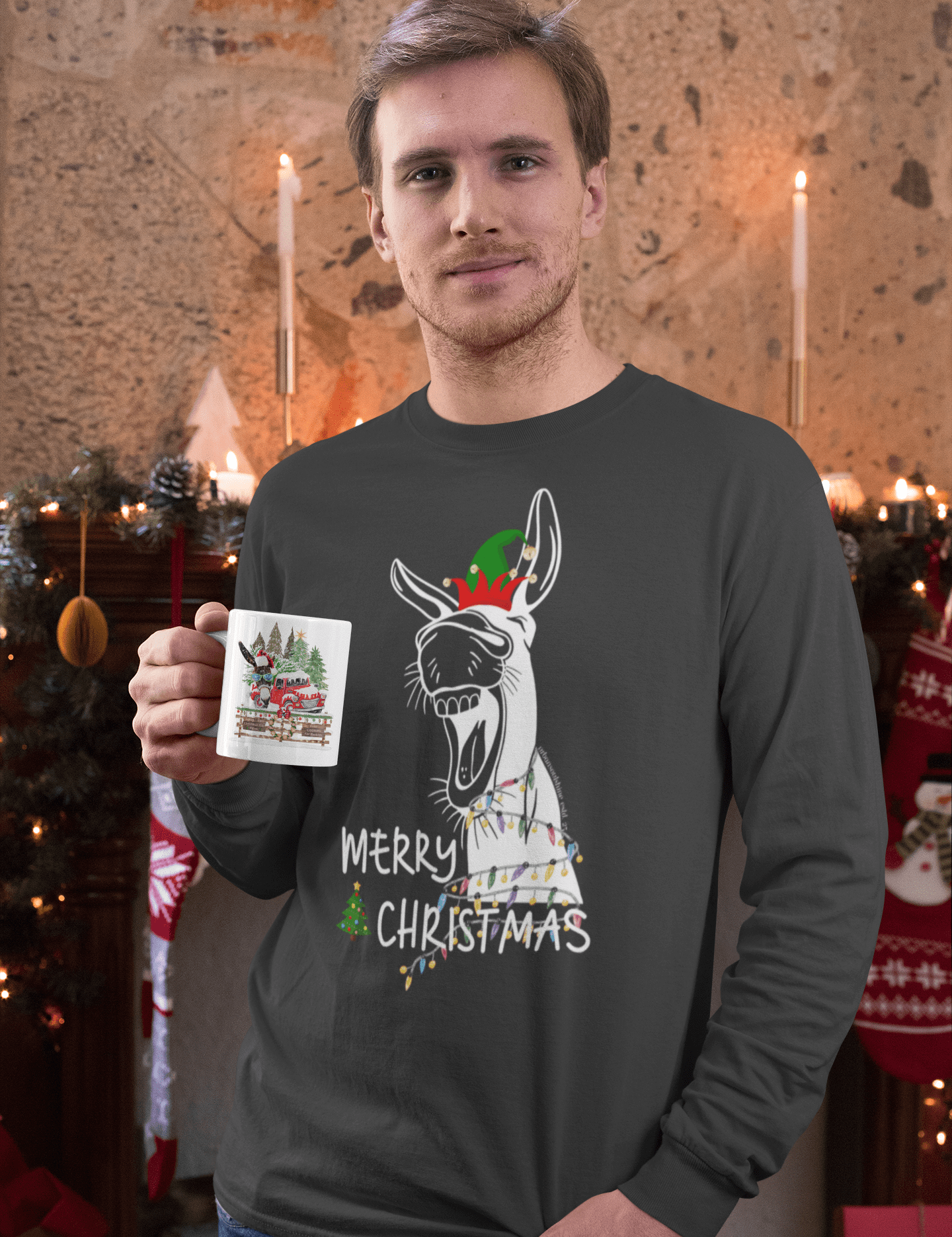 A person wearing the Merry Donkey Long Sleeve Christmas Tee by Urban SoulShine & co stands in a decorated room. They hold a mug with a similar holiday theme. In the background are candles, stockings, and evergreen garlands, all enhancing the cozy seasonal ambiance.