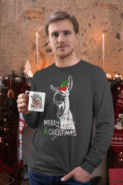 A person wearing the Merry Donkey Long Sleeve Christmas Tee by Urban SoulShine & co stands in a decorated room. They hold a mug with a similar holiday theme. In the background are candles, stockings, and evergreen garlands, all enhancing the cozy seasonal ambiance.