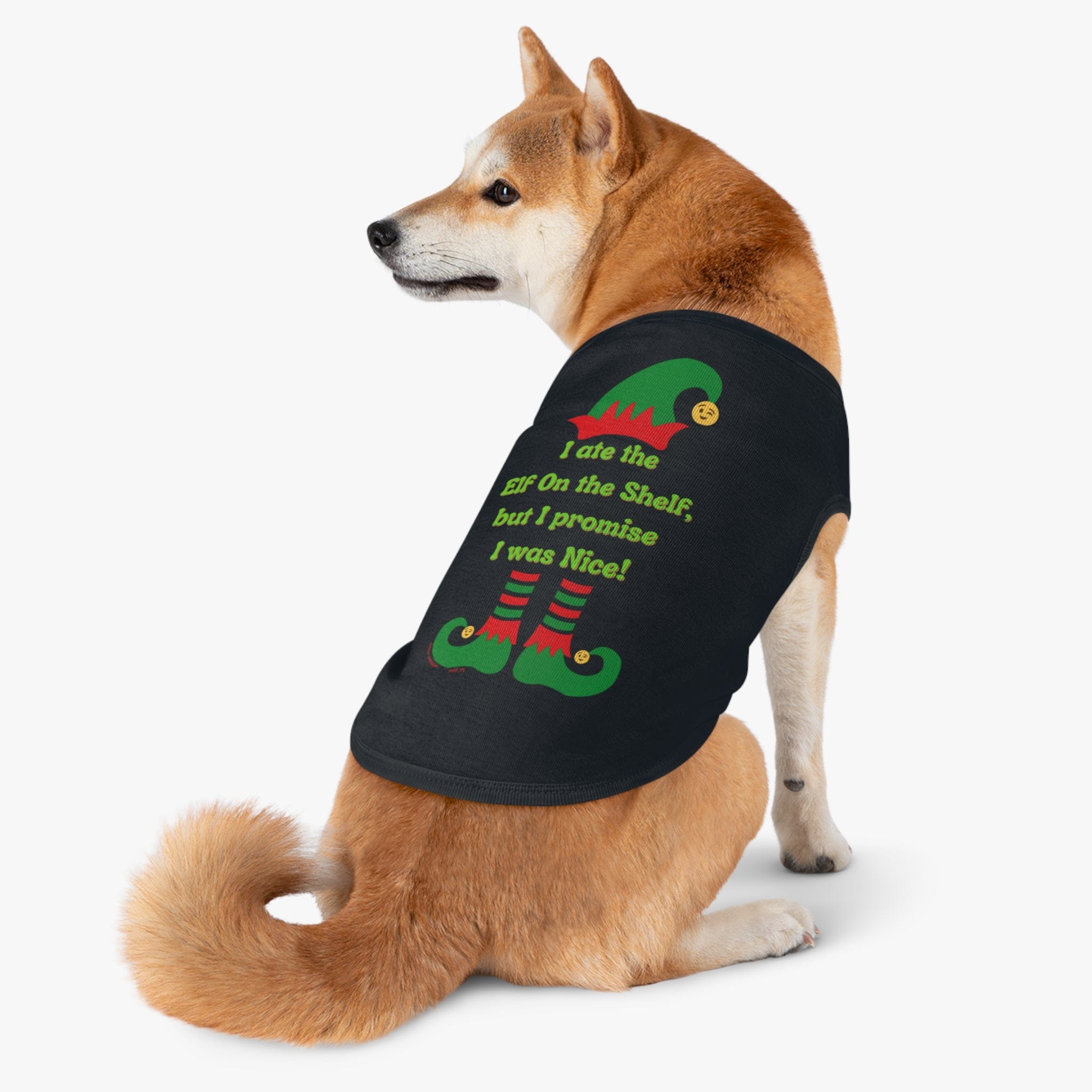 A Shiba Inu dog is dressed in the Urban SoulShine & co "I Ate the Elf On the Shelf Holiday Pet Shirt." This black shirt features a playful design of elf shoes and the humorous phrase, "I ate the Elf on the Shelf, but I promise I was nice!" It's both stylish and machine washable. The dog sits against a plain white background, looking to the side.