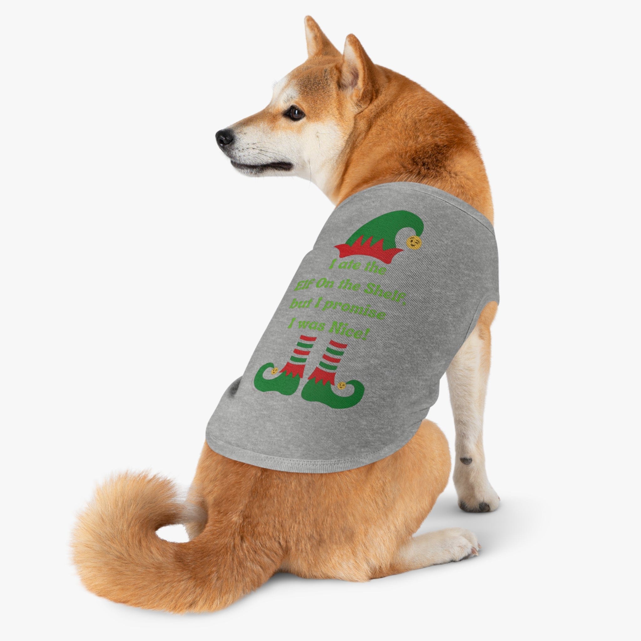 A Shiba Inu sits and looks back, wearing the Urban SoulShine & co "I Ate the Elf On the Shelf Holiday Pet Shirt," featuring a gray tone with a colorful design of elf shoes and the cheeky phrase "I ate the elf on the shelf but I promise I was nice!" This charming piece of pet apparel is machine washable, guaranteeing easy maintenance for your furry friend's wardrobe.