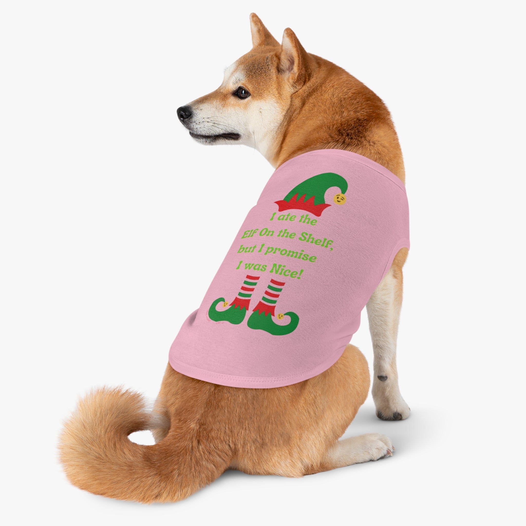 A Shiba Inu is decked out in a custom Urban SoulShine & co pet shirt, featuring a festive pink design. The "I Ate the Elf On the Shelf Holiday Pet Shirt" humorously reads, "I ate the ELF on the Shelf, but I promise I was Nice!" with images of elf legs and shoes. Suitable for machine washing, it complements the dog's curled tail as it looks over its shoulder.