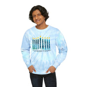 A person with medium-length dark hair is wearing the "Prosperity Love & Laughter Tie-Dye Hanukkah Sweatshirt (Unisex)" from Urban SoulShine & Co. Made from pre-shrunk cotton, this custom sweatshirt features nine lit candles and the phrase "Prosperity, Love & Laughter." They are standing against a white background, smiling with hands in pockets.