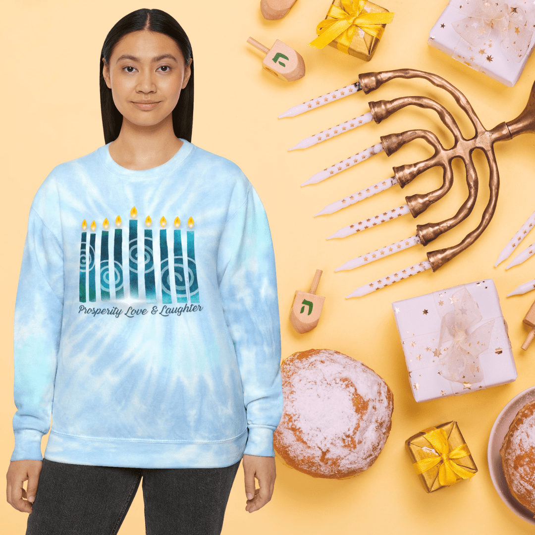 Wearing the Urban SoulShine & co Prosperity Love & Laughter Tie-Dye Hanukkah Sweatshirt, a person stands against a yellow background. The pre-shrunk cotton sweatshirt, adorned with candle graphics in blue tie-dye, captures the festive atmosphere with nearby elements like a menorah, dreidels, gifts, and sweets that reflect the custom Hanukkah design.