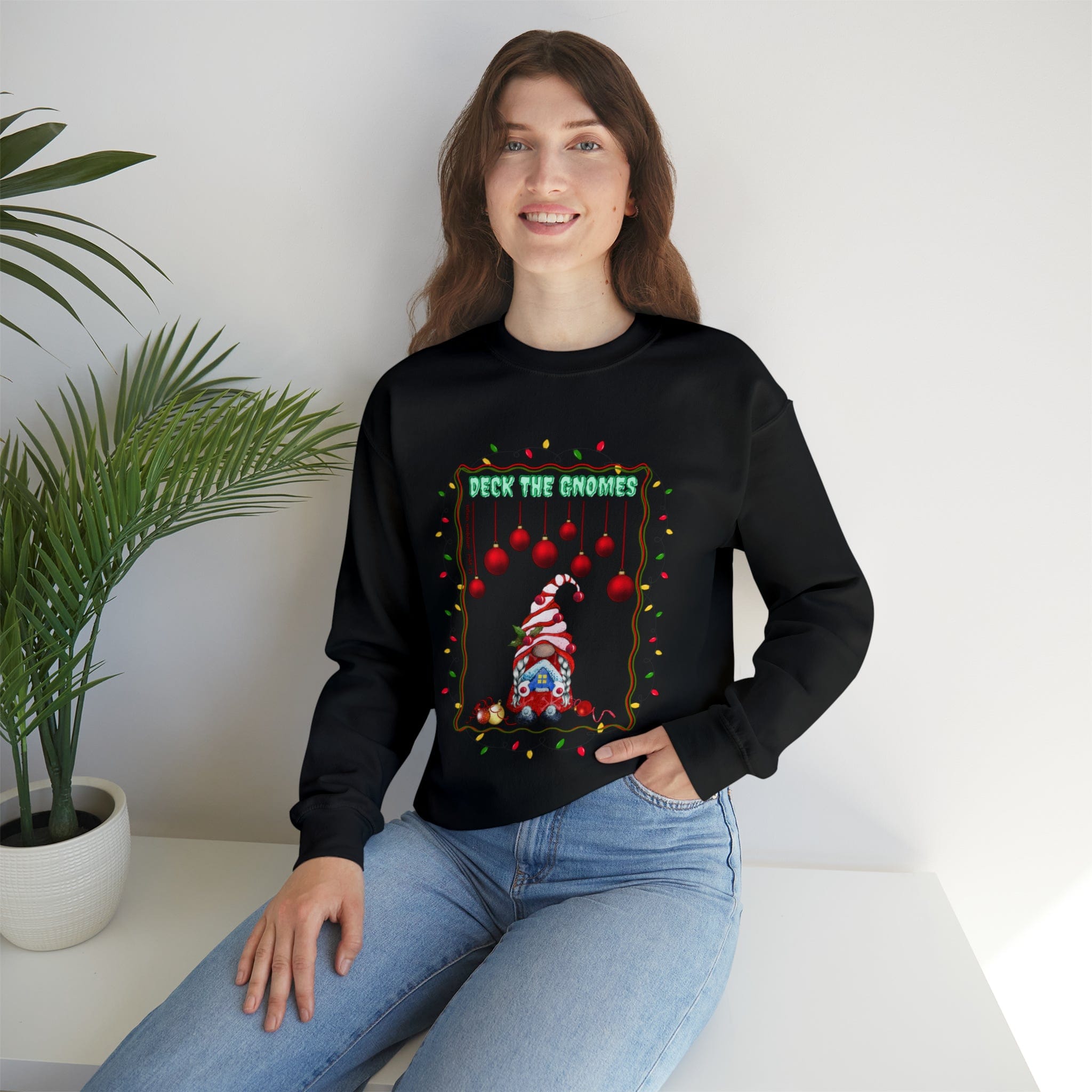 An individual with long hair is seated on a white surface, wearing Urban SoulShine & co's "Deck The Gnomes Holiday Crewneck Sweatshirt (Unisex)." This black sweater showcases a festive design with a gnome, red ornaments, and the words "DECK THE GNOMES." Made from a polyester cotton blend, this cozy crewneck spells comfort. A potted plant can be seen on the left side.