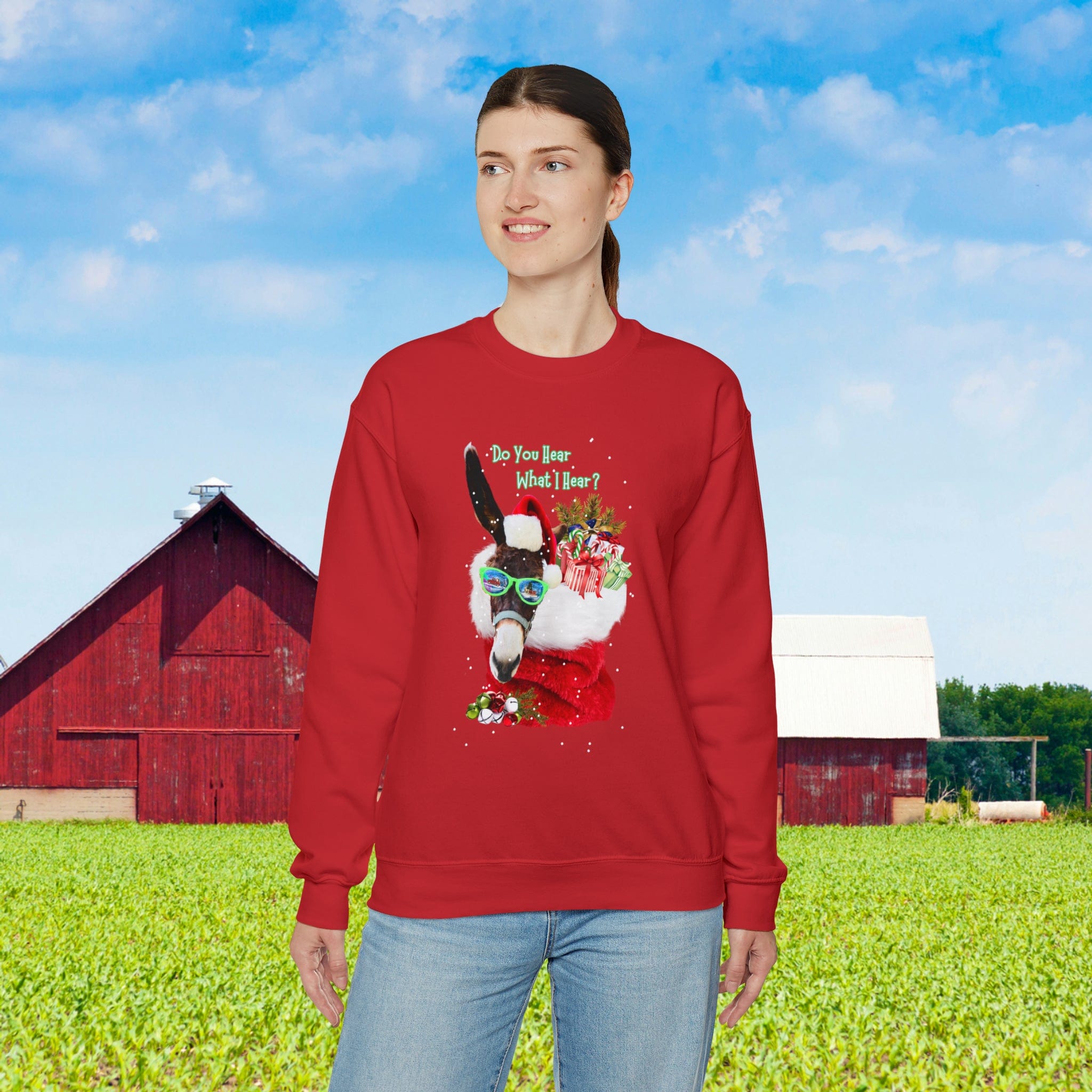 A person stands outdoors wearing the Urban SoulShine & co "Do You Hear What I Hear Donkey Claus Christmas Sweatshirt" in a vibrant red. This unisex heavy blend crewneck, featuring a festive donkey donned in a Santa hat, complements the scenic backdrop of red and white barns with a lush green field under the clear blue sky, radiating holiday cheer.