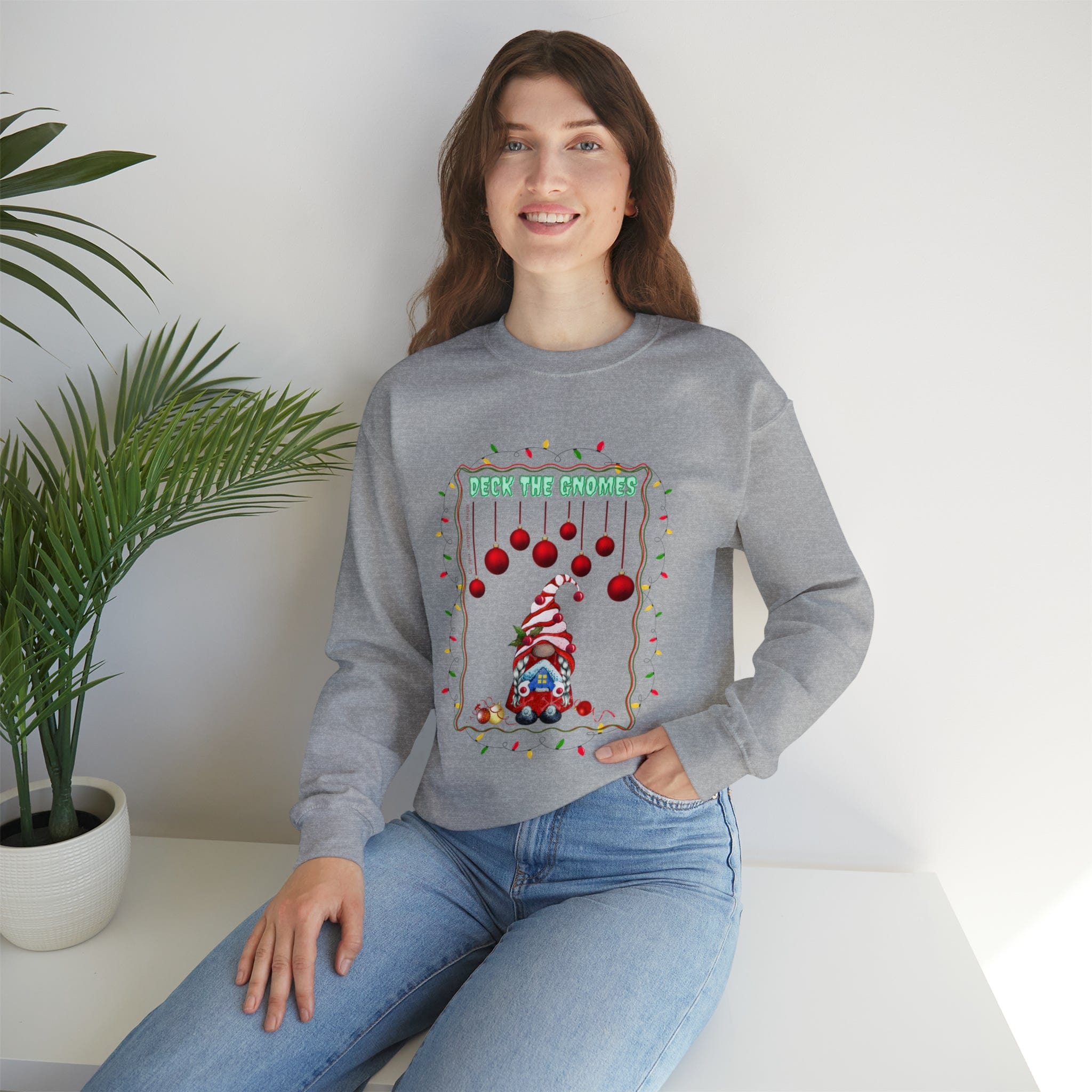 Sporting a warm smile, a person with long brown hair sits on a white surface beside a vibrant green plant, donning Urban SoulShine & co's Deck The Gnomes Holiday Crewneck Sweatshirt. This cozy gray unisex piece, adorned with charming gnomes and ornaments alongside the "Deck the Gnomes" text, pairs perfectly with their light blue jeans and offers unbeatable comfort in its polyester cotton blend.