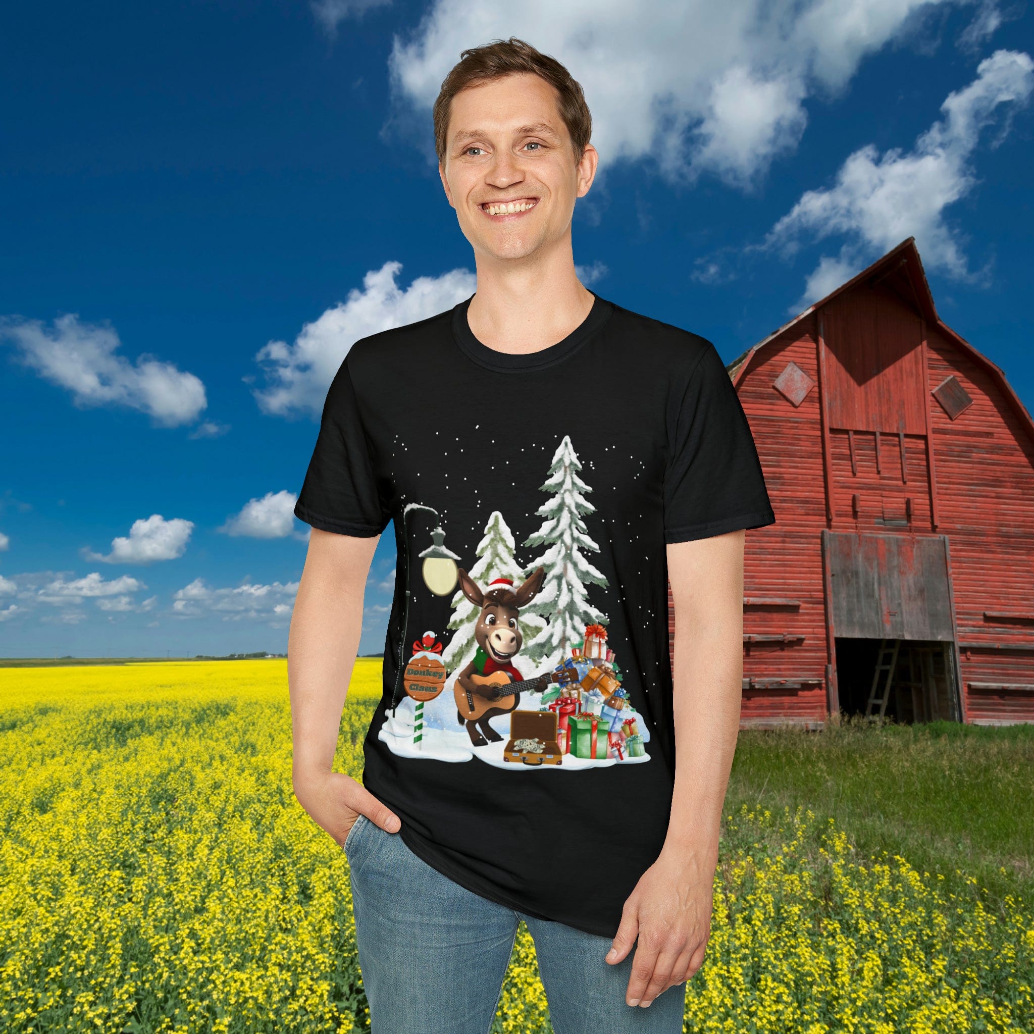 A person stands in a field of yellow flowers in front of a red barn, wearing the festive Donkey Claus Street Singer Holiday Soft Tee (Unisex) from Urban SoulShine & co. The black T-shirt features reindeer, snow-covered trees, and various gifts and is made from 100% cotton. Above, the sky is clear and blue.