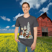 Against the backdrop of a red barn and a field of yellow flowers, someone is wearing the Donkey Claus Street Singer Holiday Soft Tee (Unisex) from Urban SoulShine & co. The gray T-shirt is crafted from 100% cotton and showcases a festive cartoon moose holding a guitar surrounded by evergreen trees, gifts, a mailbox, and a streetlamp in a snowy setting.