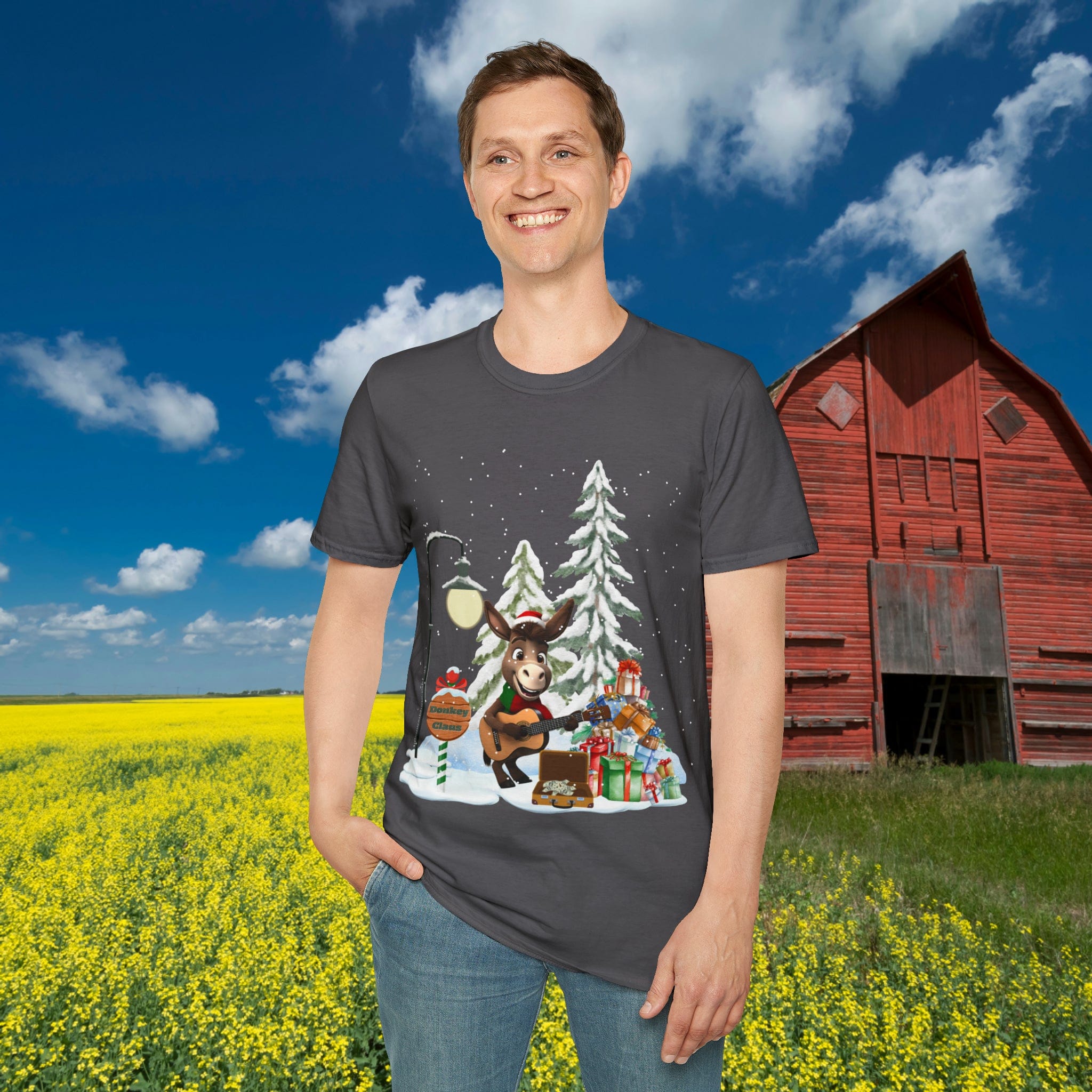 Against the backdrop of a red barn and a field of yellow flowers, someone is wearing the Donkey Claus Street Singer Holiday Soft Tee (Unisex) from Urban SoulShine & co. The gray T-shirt is crafted from 100% cotton and showcases a festive cartoon moose holding a guitar surrounded by evergreen trees, gifts, a mailbox, and a streetlamp in a snowy setting.