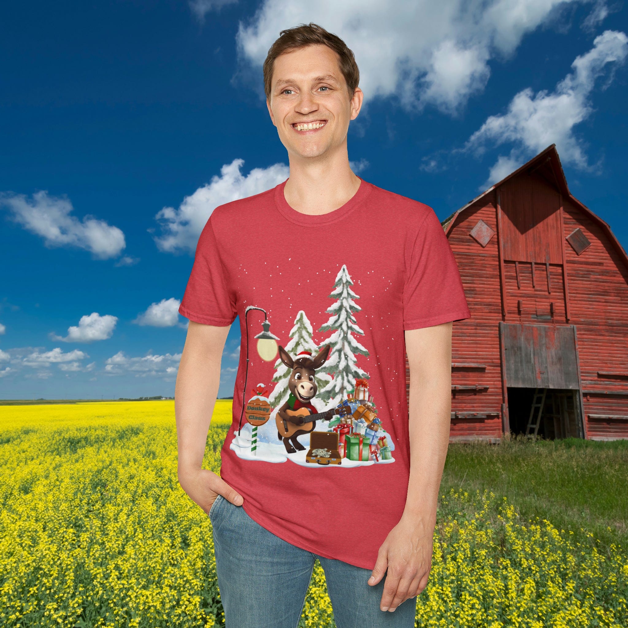 In a field of yellow flowers, a man stands smiling while wearing the Donkey Claus Street Singer Holiday Soft Tee by Urban SoulShine & co. This 100% cotton red T-shirt features a cartoon reindeer playing guitar next to snow-covered trees and Christmas presents. In the background, there's a red barn under a blue sky with some clouds.