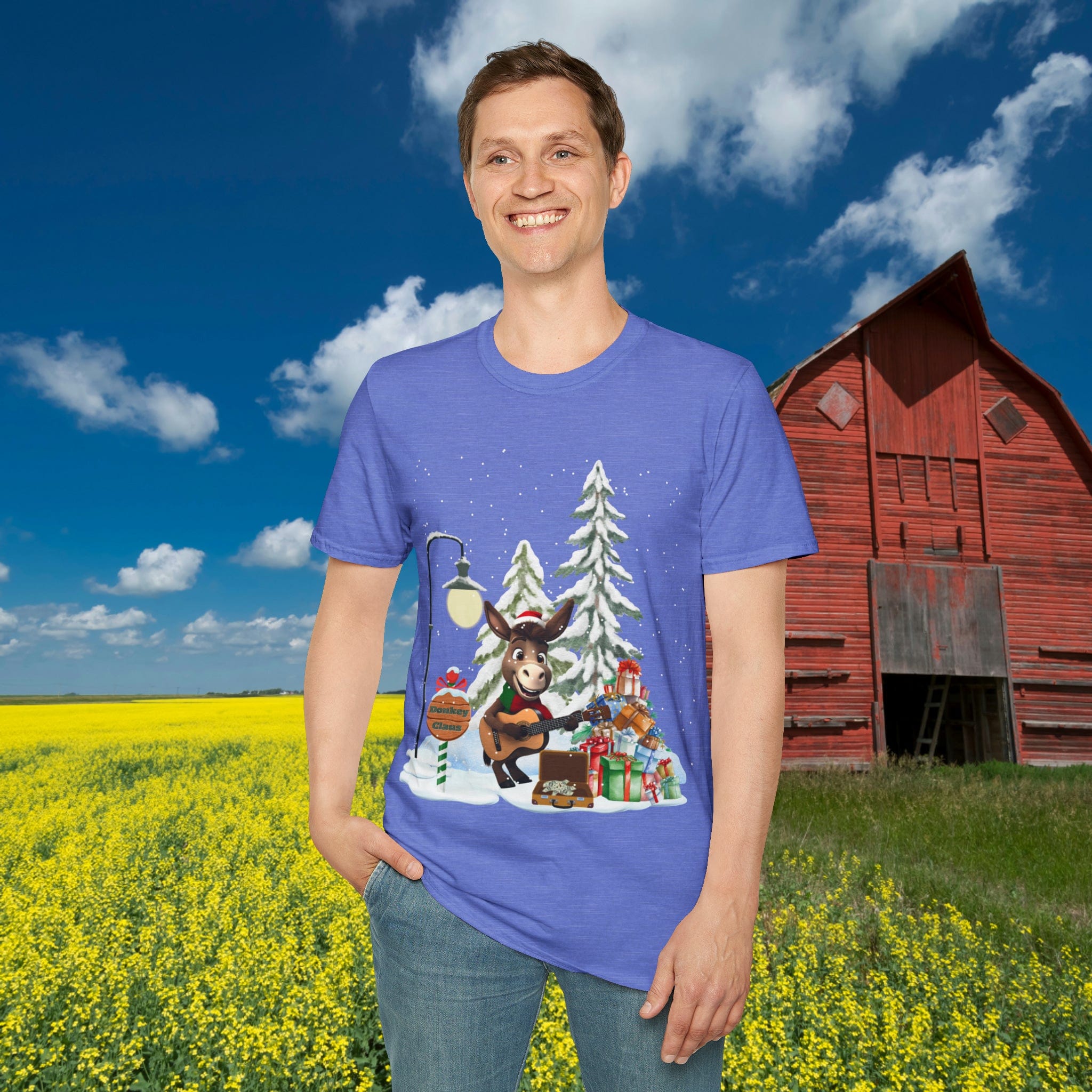 In a picturesque scene, someone smiles amidst a field of yellow flowers while wearing the Donkey Claus Street Singer Holiday Soft Tee by Urban SoulShine & co. This 100% cotton blue T-shirt features an adorable cartoon moose with a Christmas-themed design. A rustic red barn and a blue sky dotted with clouds add the perfect backdrop.