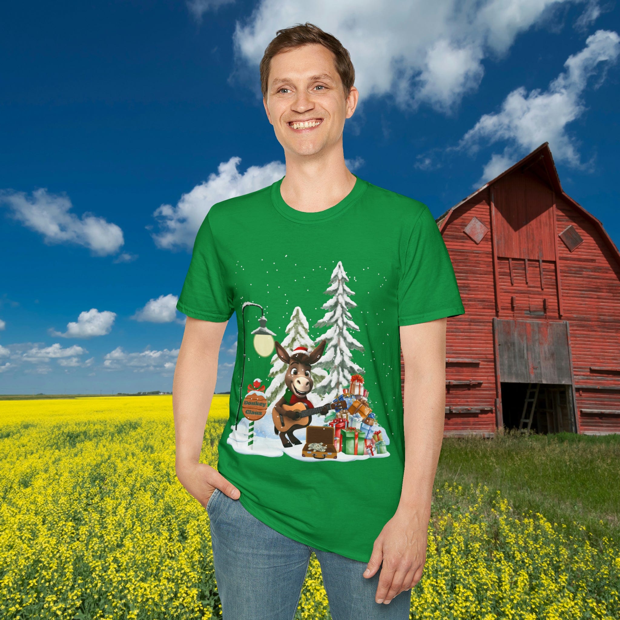 In a field of yellow flowers under a blue sky, a person stands smiling while wearing the Urban SoulShine & co Donkey Claus Street Singer Holiday Soft Tee, showcasing a festive design of Donkey Claus near snow-covered trees and presents. This unisex t-shirt is made of 100% cotton and beautifully complements the scene, with a red barn visible in the background.