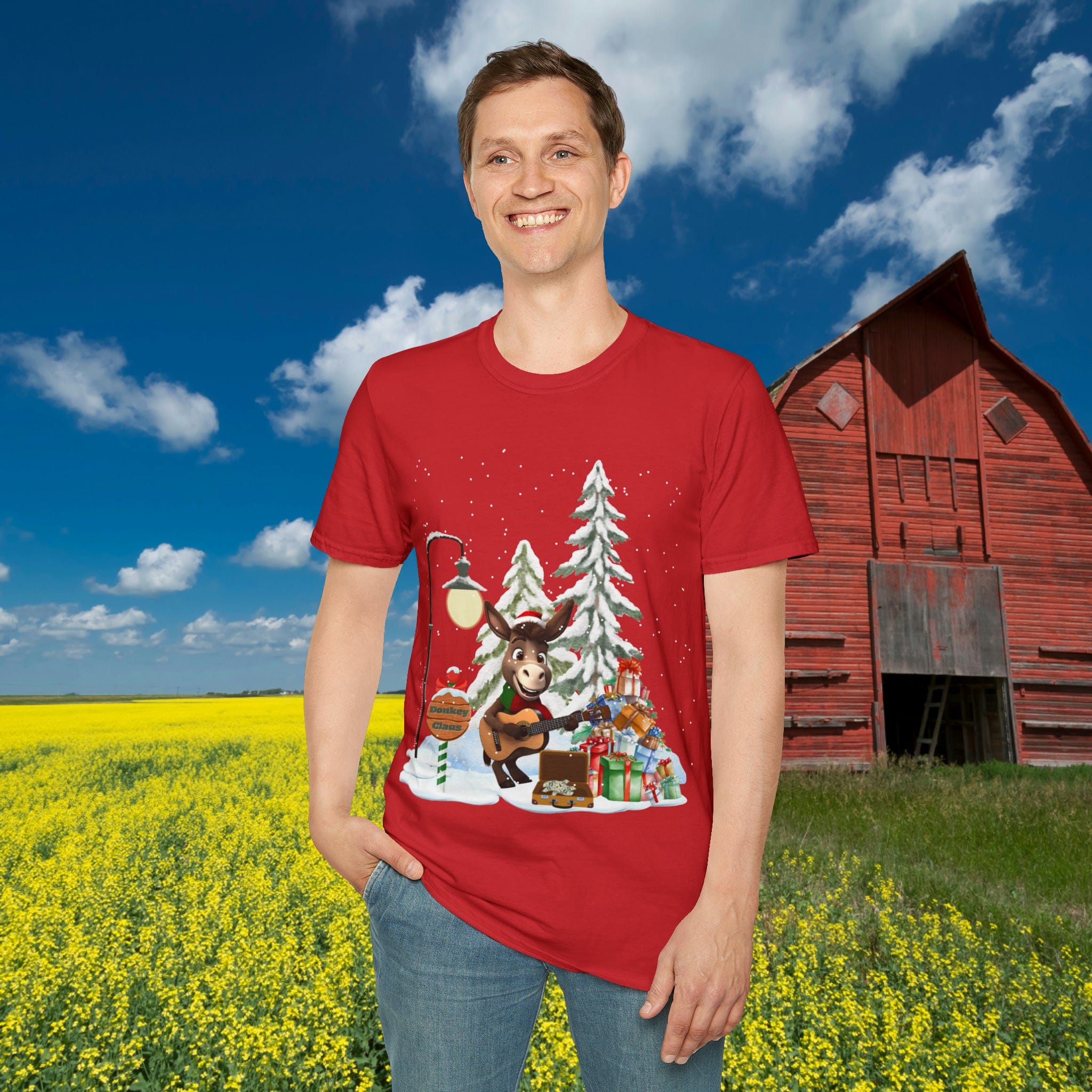 A person stands in a field of yellow flowers wearing the Urban SoulShine & co Donkey Claus Street Singer Holiday Soft Tee, a 100% cotton, unisex t-shirt featuring a festive design with a donkey playing guitar amidst snowy trees and gifts. A red barn is in the background under a blue sky with clouds.