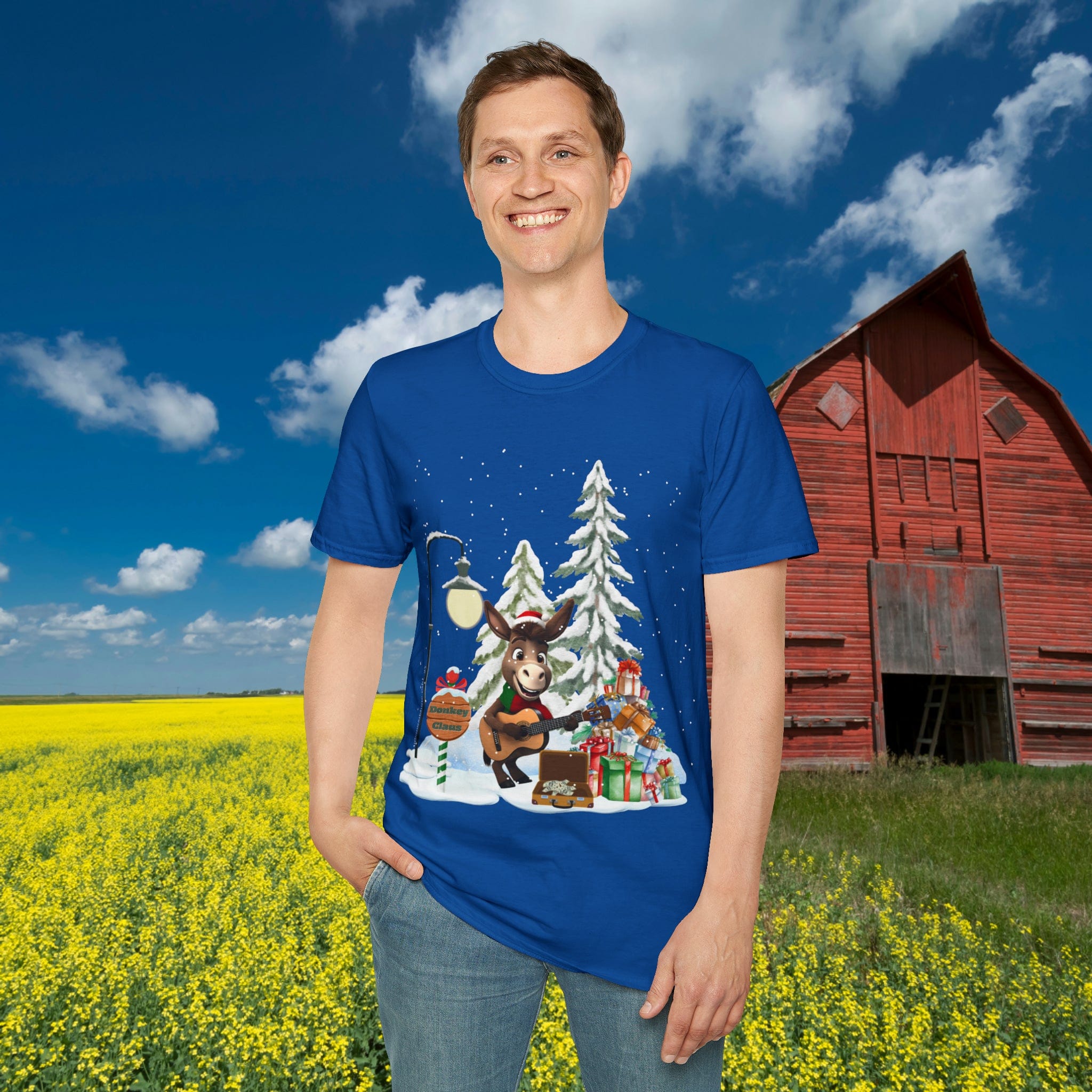 A smiling person stands in a field of yellow flowers, wearing the festive Donkey Claus Street Singer Holiday Soft Tee (Unisex) from Urban SoulShine & co, made of 100% cotton. The design features a moose, snow-covered trees, and gifts. A red barn and a blue sky with clouds serve as the backdrop, enhancing the cheerful scene.