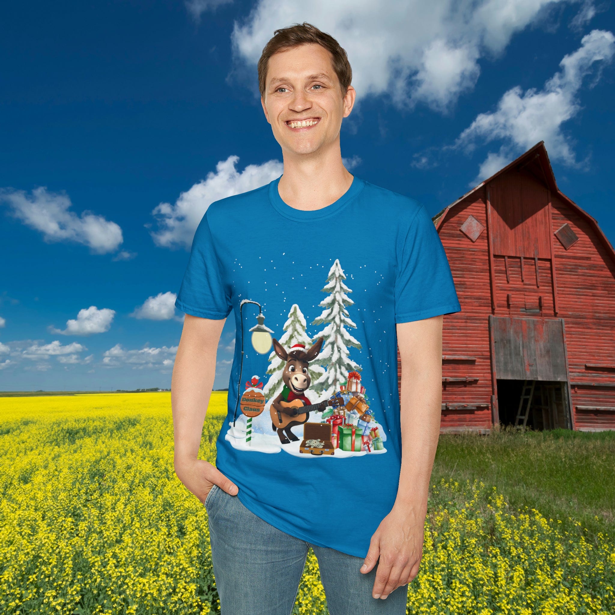 In a field of yellow flowers, someone smiles while wearing the Urban SoulShine & co's Donkey Claus Street Singer Holiday Soft Tee, made from 100% cotton. The unisex blue t-shirt boasts a festive reindeer design and embodies casual charm with one hand casually tucked in their pocket. A red barn stands in the background under a sky dotted with clouds.